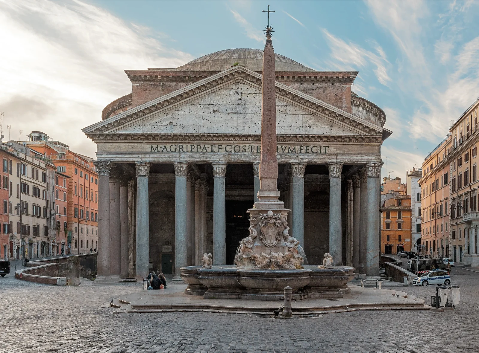 The Pantheon in Rome seen from the piazza: a columned portico with a triangular pediment and Latin inscription, the low dome rising behind it, and an obelisk fountain in front